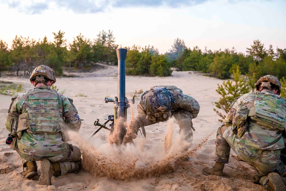 A soldier fires a weapon as two other soldiers kneel beside.