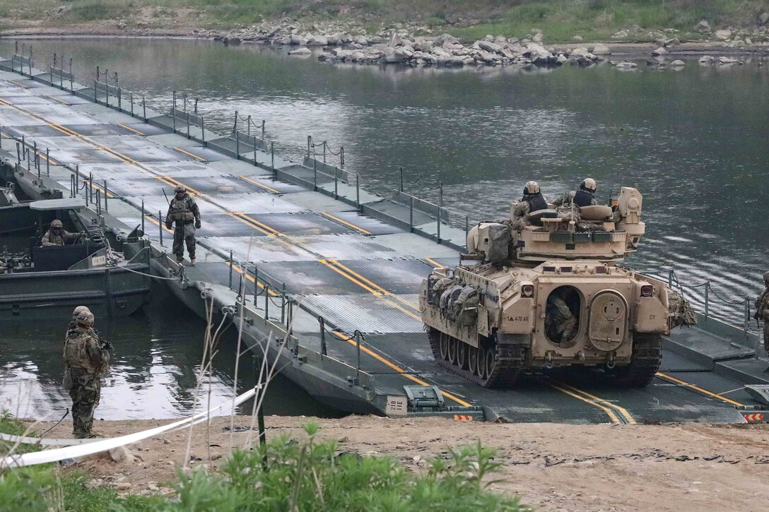 A tank with several soldiers crosses a bridge while other soldiers look on.