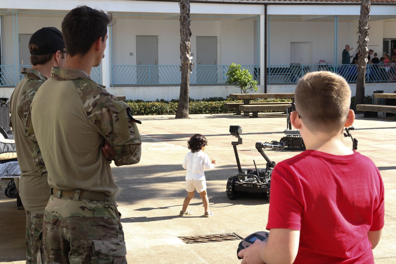 Two uniformed service members stand aside two children interacting with remote controlled devices.