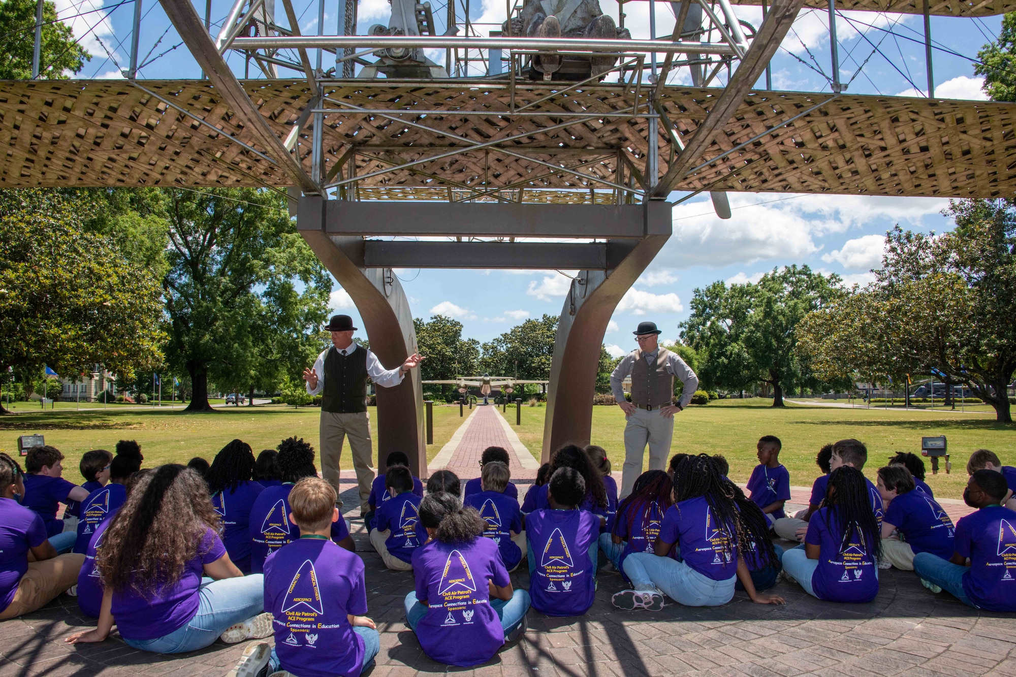 Maxwell AFB, Ala. – Elementary students from Bear Exploration Center For Mathematics, Science and Technology School perch in the shade of the Wright Flyer replica as they learn about the history of aviation from “Orville” and “Wilbur Wright,” May 13, 2022. (US Air Force photo by Melanie Rodgers Cox/Released)