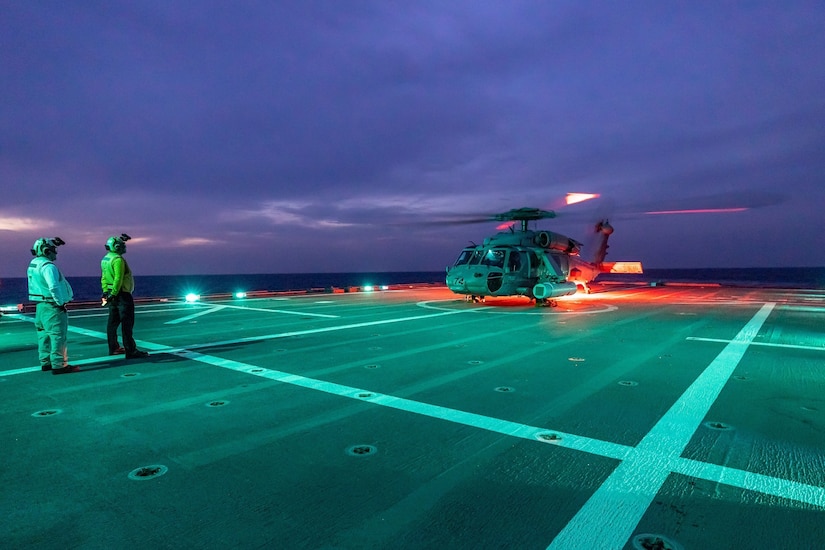 A helicopter sits on a ship lit up against a dark sky.