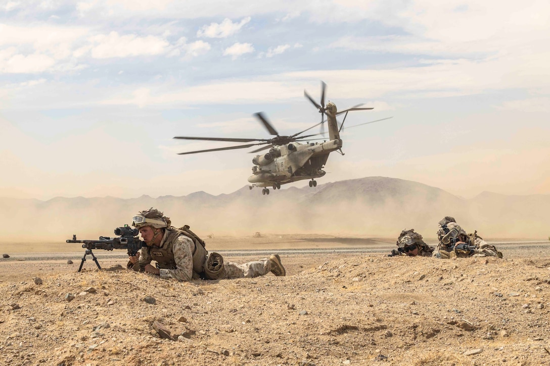 Three Marines lie on the ground holding weapons as a helicopter flies in the background.