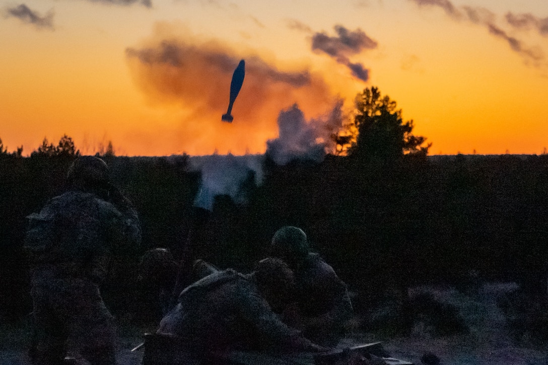 Soldiers fire a weapon during training under a sunlit sky.