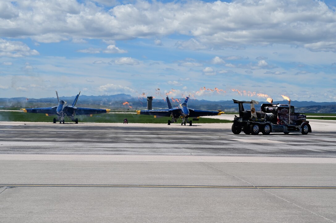 The Shockwave Jet Truck drives past two U.S. Navy Flight Demonstration Squadron Blue Angels at the 2022 Ellsworth Air and Space Show on Ellsworth Air Force Base, S.D., May 15, 2022. The Shockwave is a custom-built race truck featuring three jet engines with 36,000 horsepower. (U.S. Air Force photo by Staff Sgt. Hannah Malone)