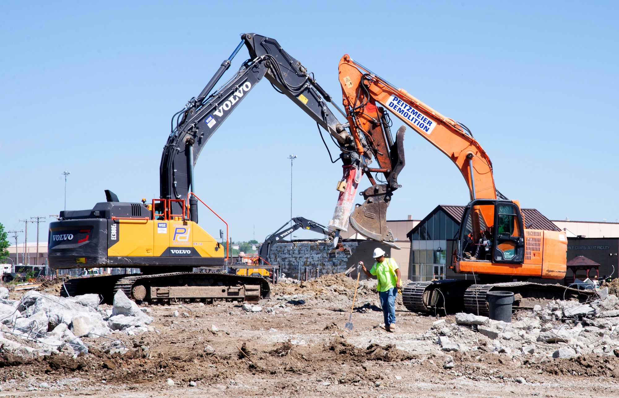 A man looks on as two large trucks move dirt and an old building