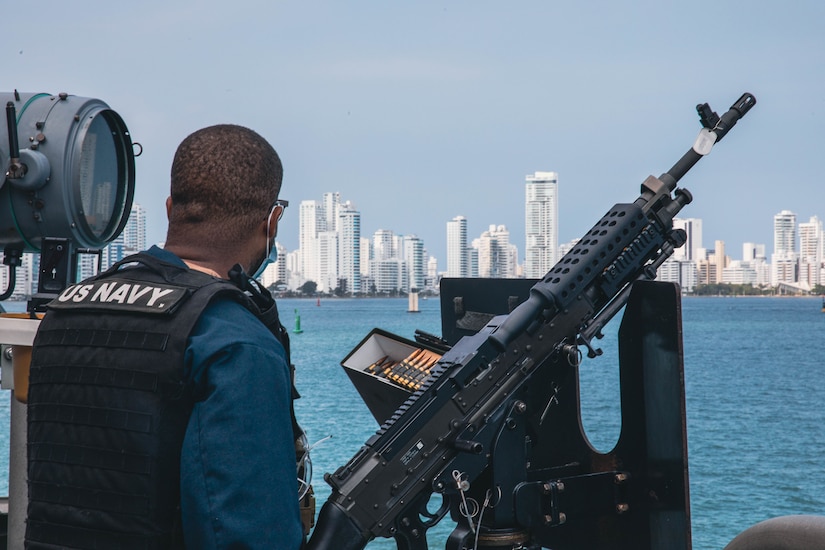 A sailor mans a gun on ship near a city’s coastline.