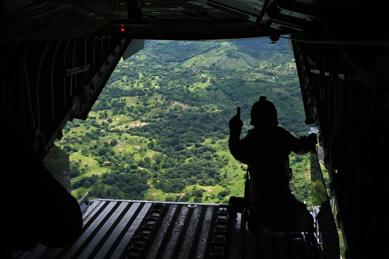 A man holds up a finger while sitting in the open back hatch of a helicopter that’s flying over a forested area.
