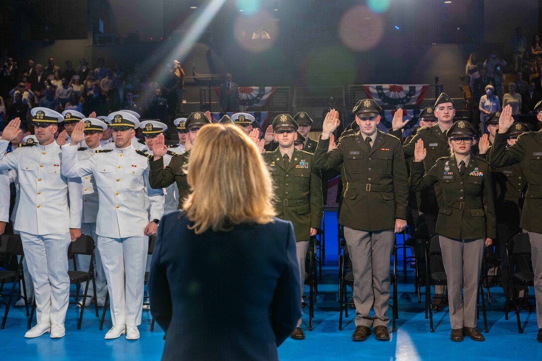 A group of military personnel in dress uniform all stand with their right hands raised. A woman stands in the foreground and looks on.
