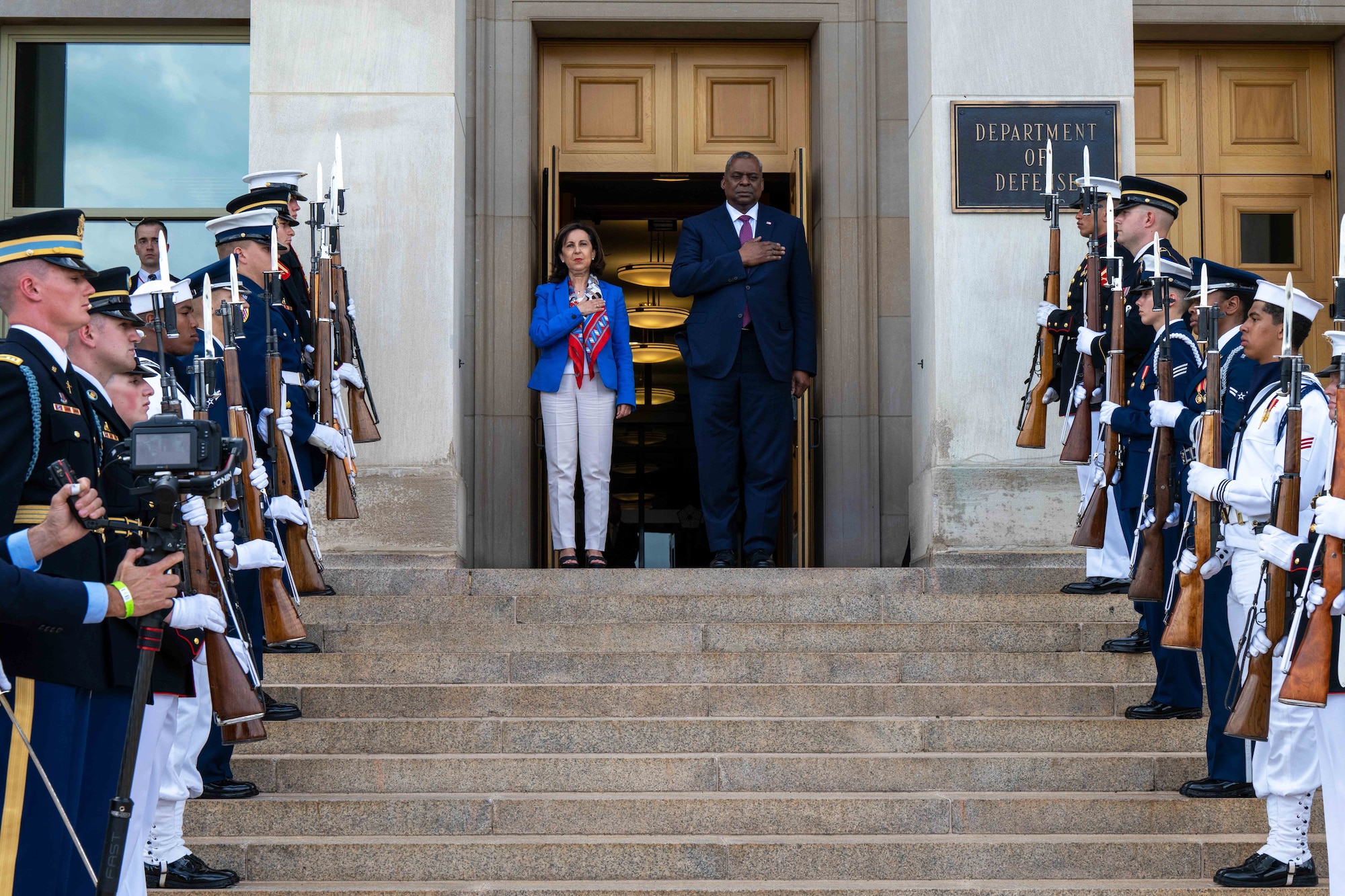 Secretary of Defense Lloyd J. Austin III stands on the steps of the pentagon with another leader, flanked by military members.