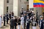 Two men stand at the top of a building’s steps as uniformed personnel stand in formation.