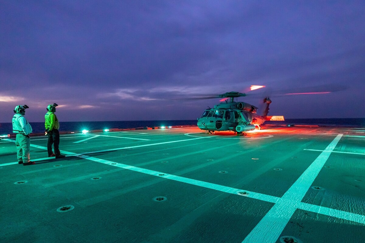 A helicopter sits on a ship lit up against a dark sky.