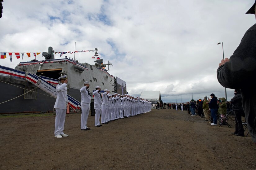 Sailors in a row salute while standing in front of a moored ship.