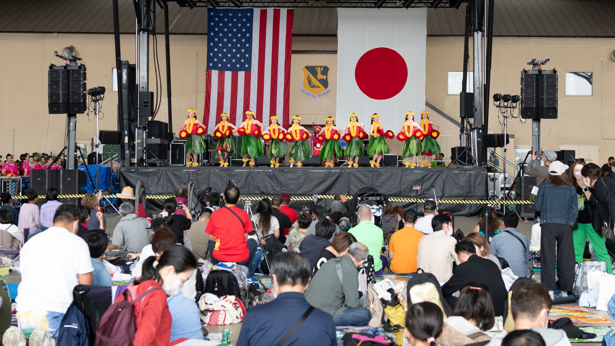 A dozen hula dancers perform on stage in front of a crowd