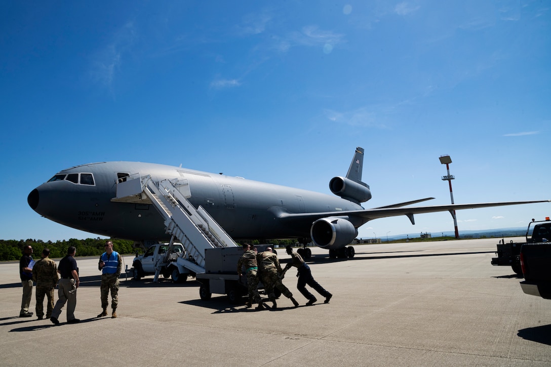 Several uniformed service members push equipment toward an aircraft with a stair car’s steps leading to its open door.