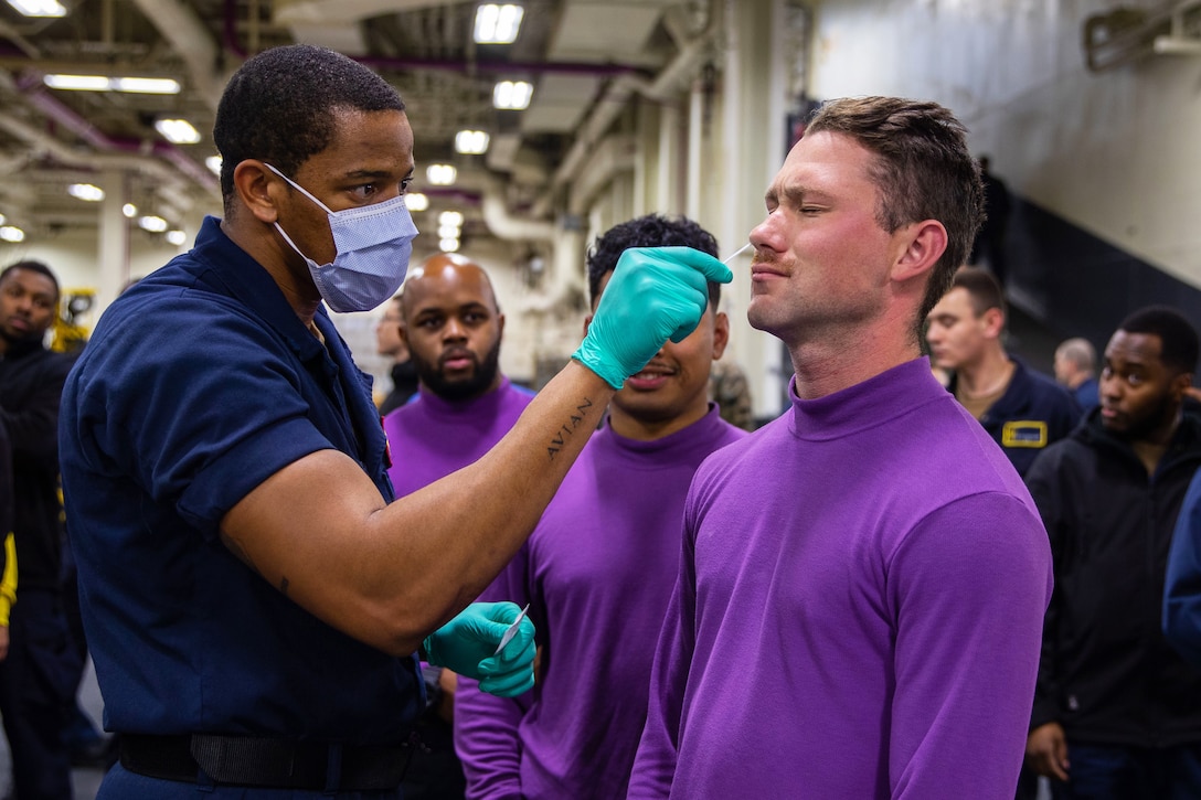 Amid crowd of service members, a uniformed man wearing a face mask and gloves swabs the nasal of a man wearing a purple turtleneck shirt.