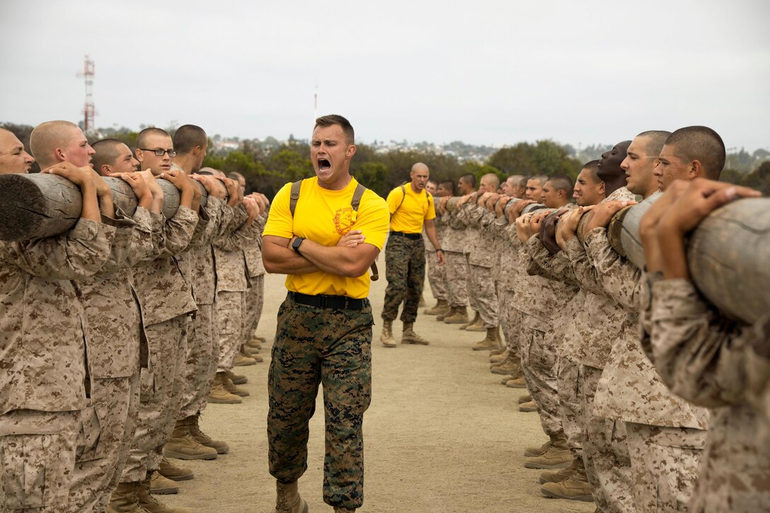 A Marine shouts at Marine Corps recruits standing in a parallel line holding logs.