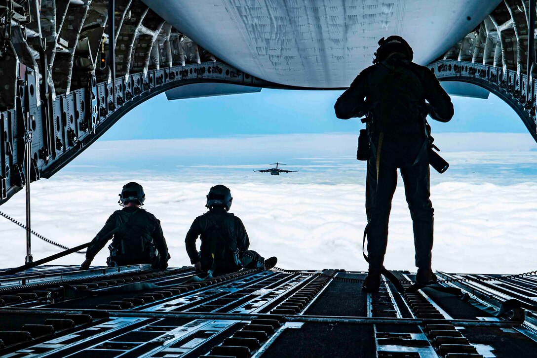 An airman stands next to two airmen sitting on a ramp of an airborne aircraft as another aircraft flies in the background.