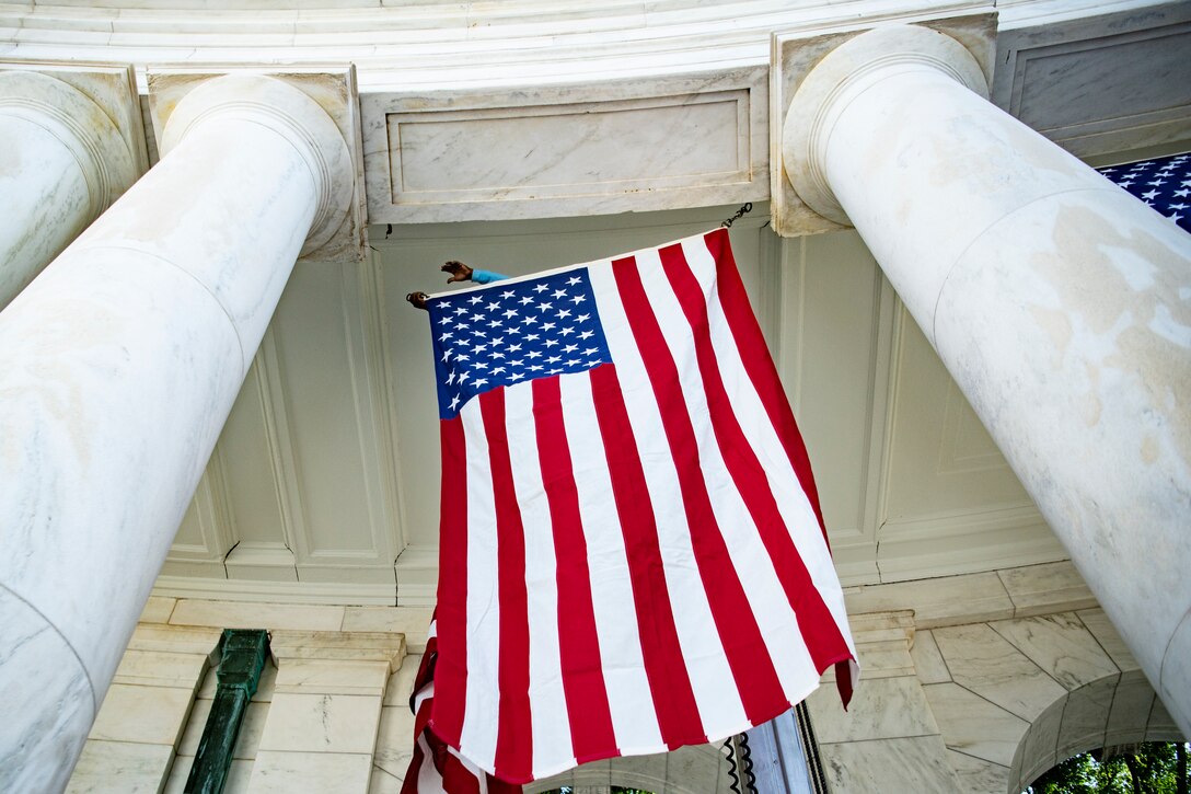 A worker reaches an American flag from one white marble column toward another to hang it.
