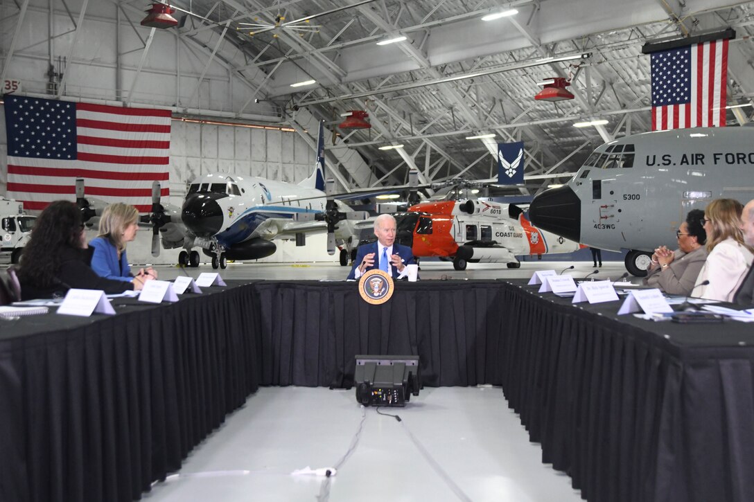 President Biden sits center at a u-shaped table as people flank him. Three aircraft can be seen behind him.