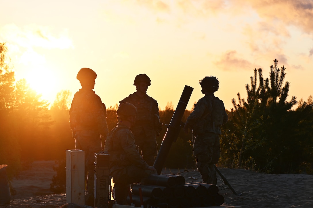 Four service members look at a weapon in a sandy area.