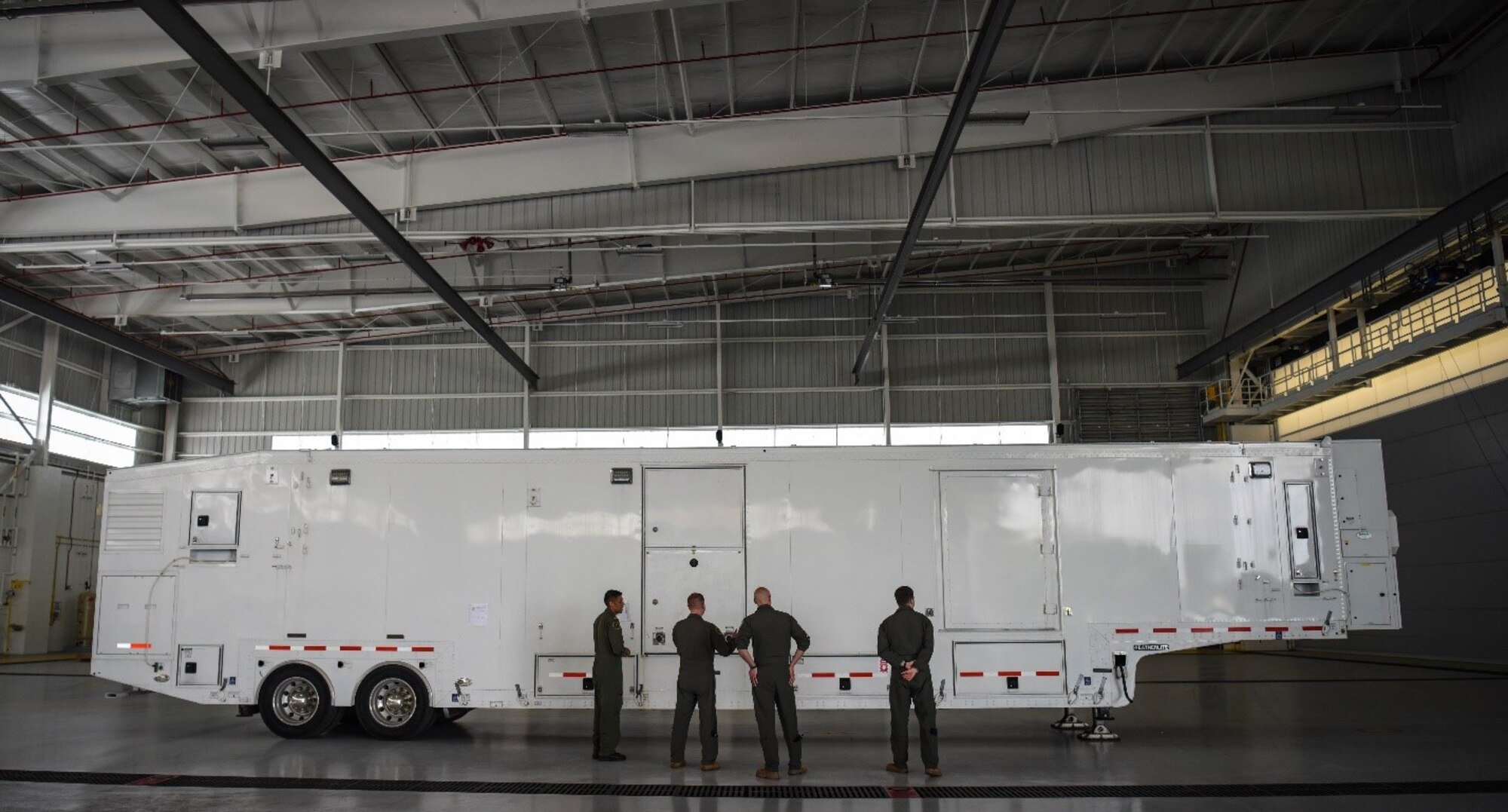 Four men stand in front of a large trailer in side a hanger.