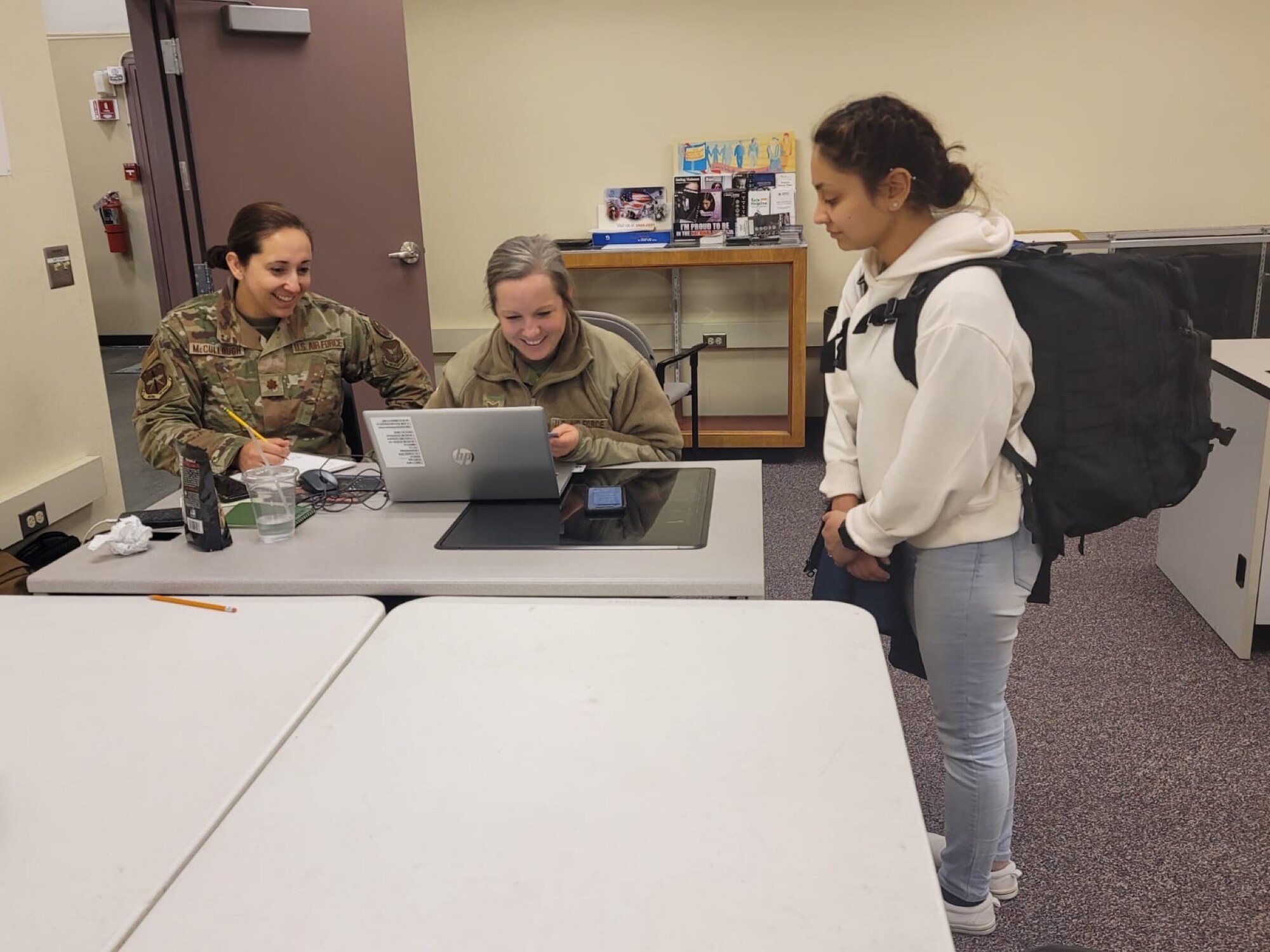 Two uniformed Airmen sit behind a table, one on a laptop, while an Airman in plain clothes stands to the right.