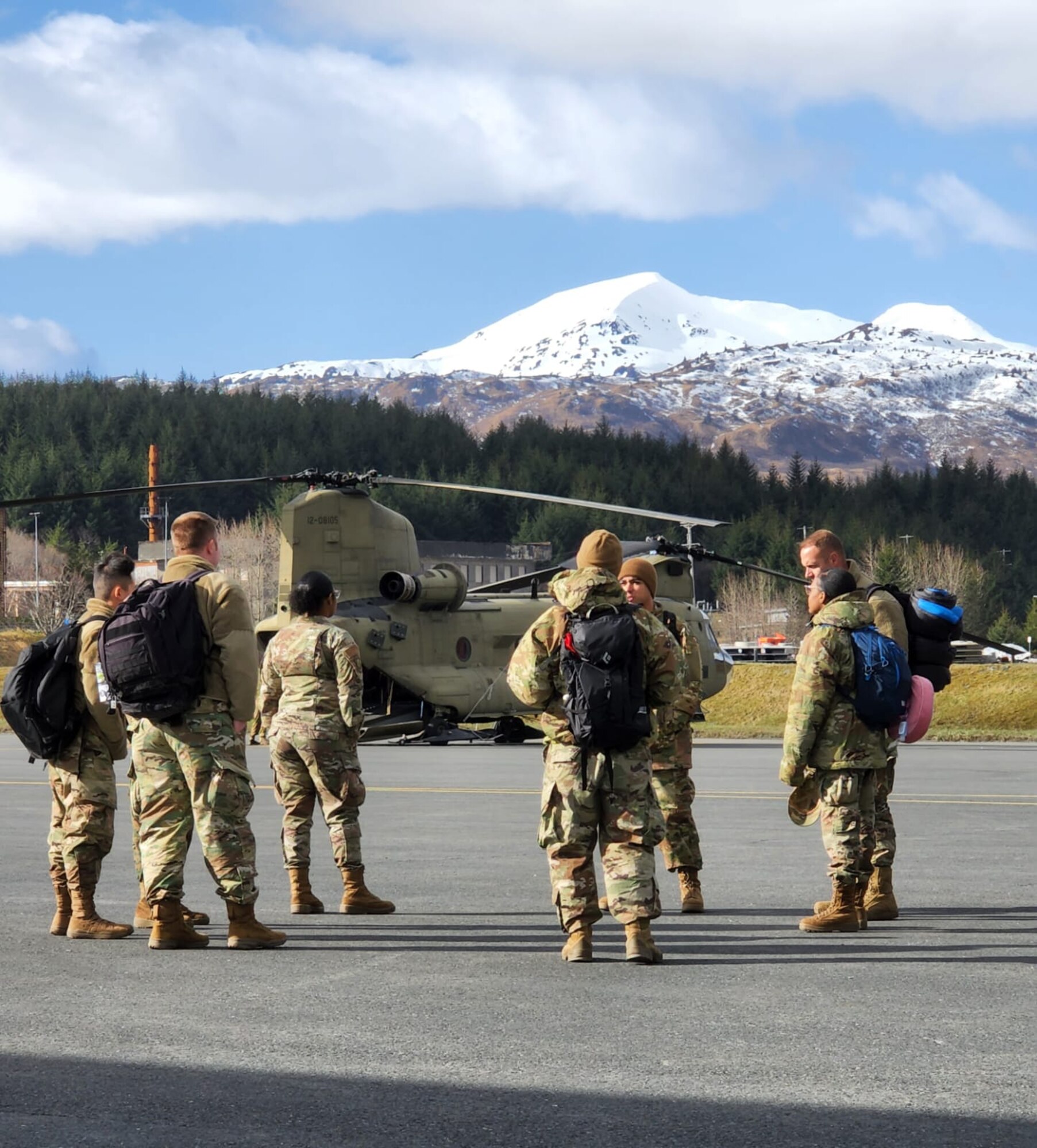 Airmen stand in the foreground, with a Chinook behind them and a snow-capped mountain in the background