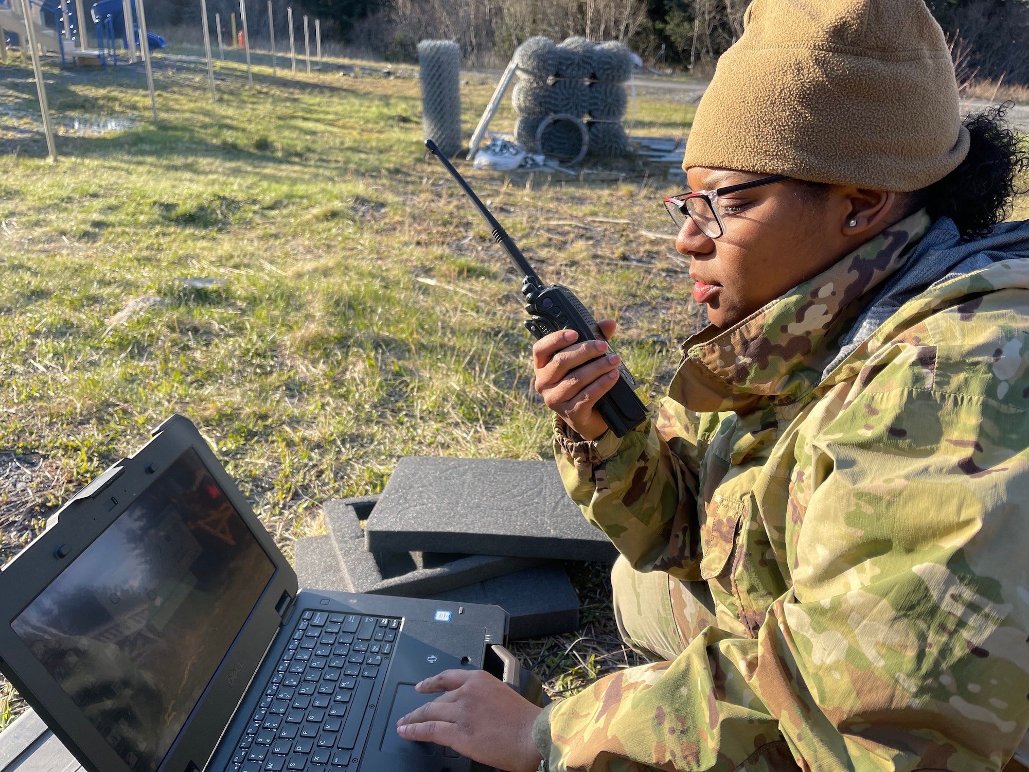 Preatto is sitting on the ground and has one hand on the trackpad of a laptop and is holding a radio in the other hand.