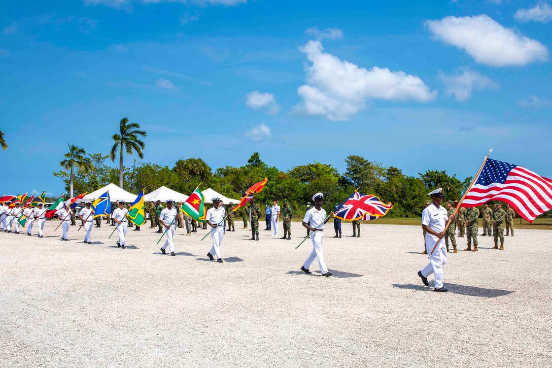 A group of service members walk in a line on a beach holding flags.