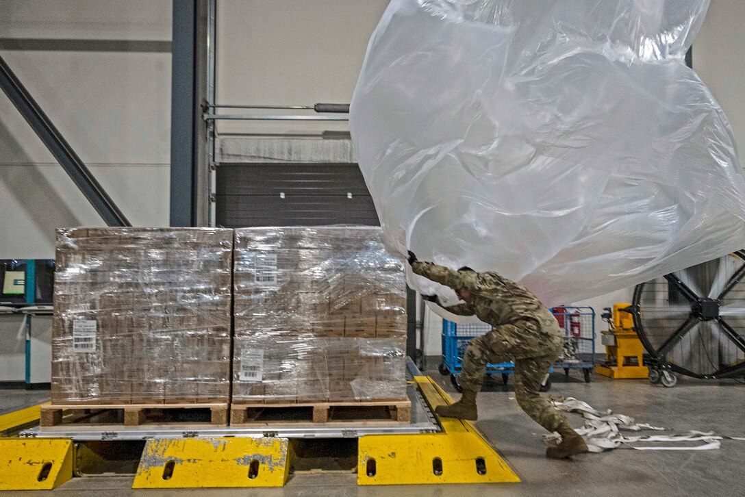 An airman maneuvers a giant plastic bag over a pallet of supplies in a warehouse-type space.