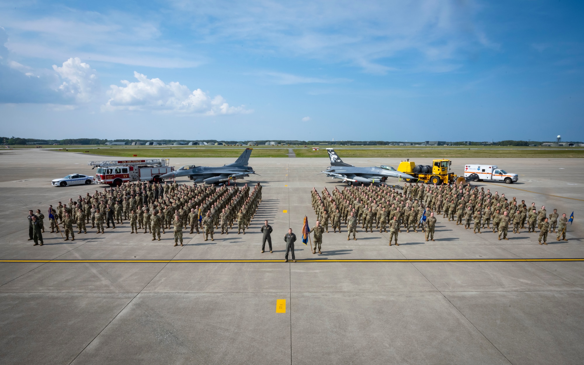Group of military members in uniform pose in front of military vehicles and aircraft for a photo.
