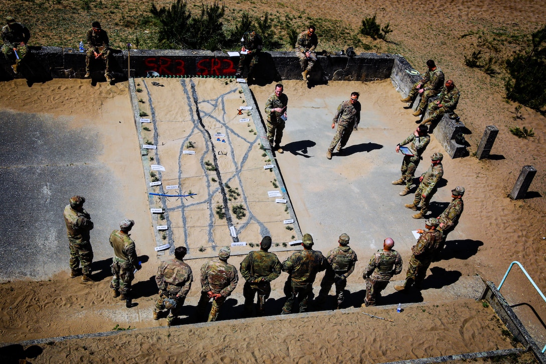 Two high-ranking U.S. service members conduct a debrief with multiple German soldiers on a concrete slab in a dirt field.