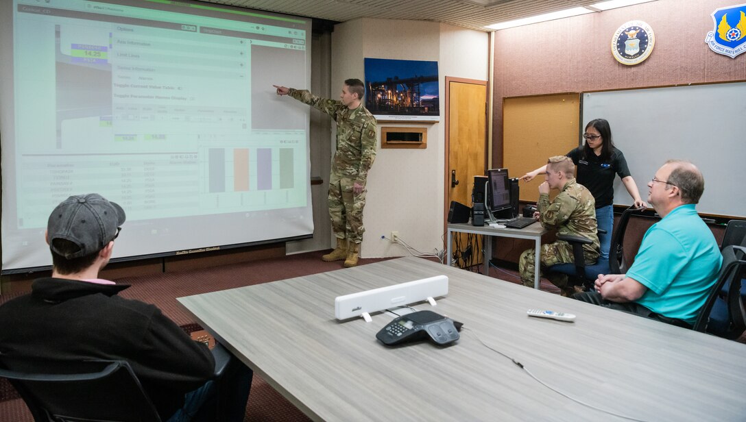Airman pointing to screen as two other people look and two other people work at a computer