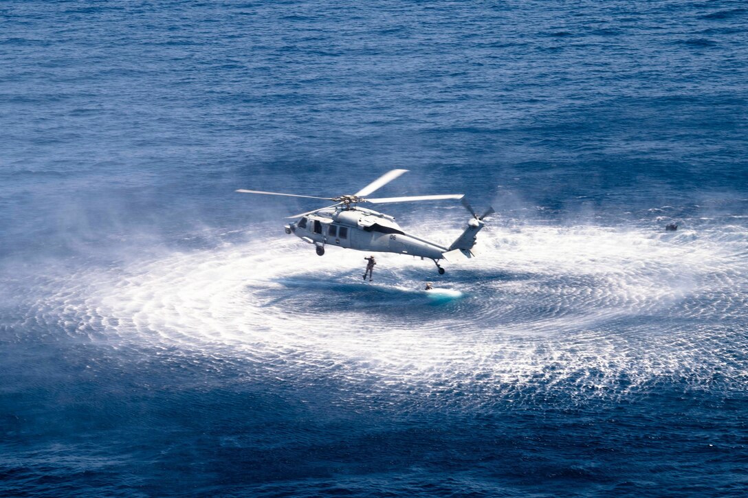 A sailor jumps from an airborne helicopter into a body of water where a fellow sailor is floating.