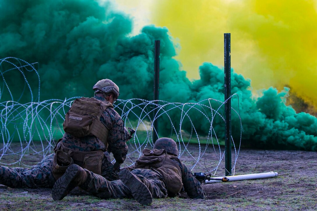 Marines prepare a torpedo in front of barbed wire as green and yellow smoke rises in the background.