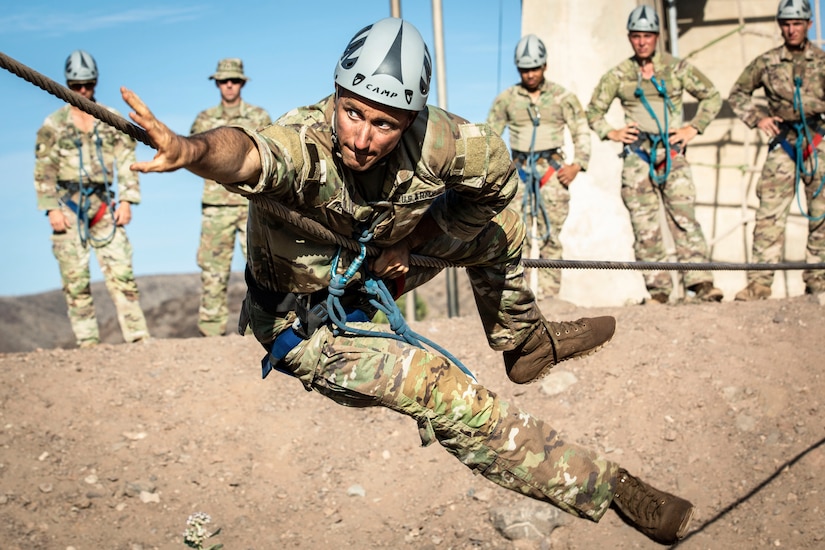 A service member climbs across rope  as fellow service members watch.