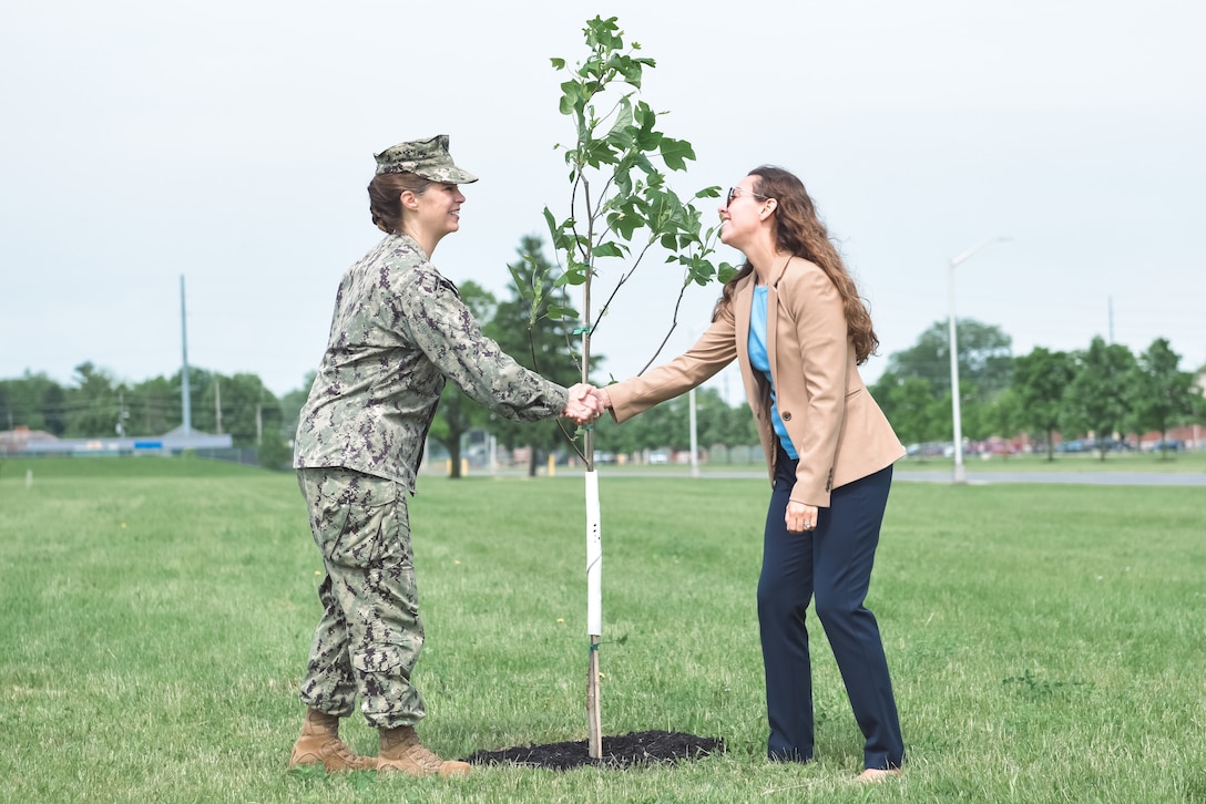 A woman in military uniform shakes the hand of another woman in a business suit outside in front of a tulip tree.