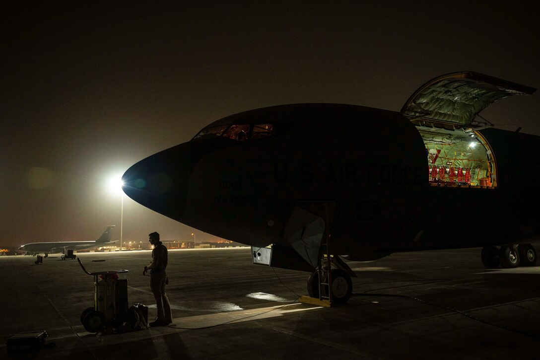 An aircraft with an open door sits on the tarmac in the dark as service member stands nearby.