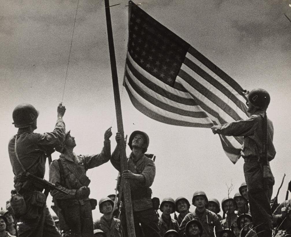 Four service members raise a U.S. flag on a flagpole while others watch in the background.