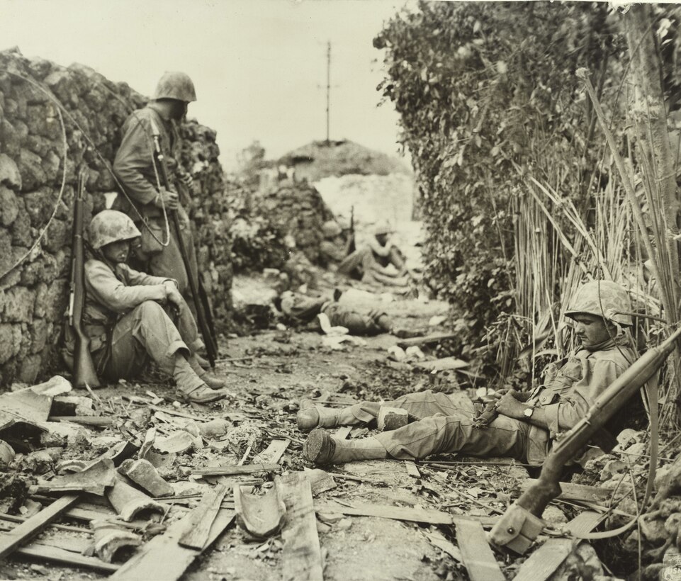 Service members relax behind a rock wall amid debris from war.