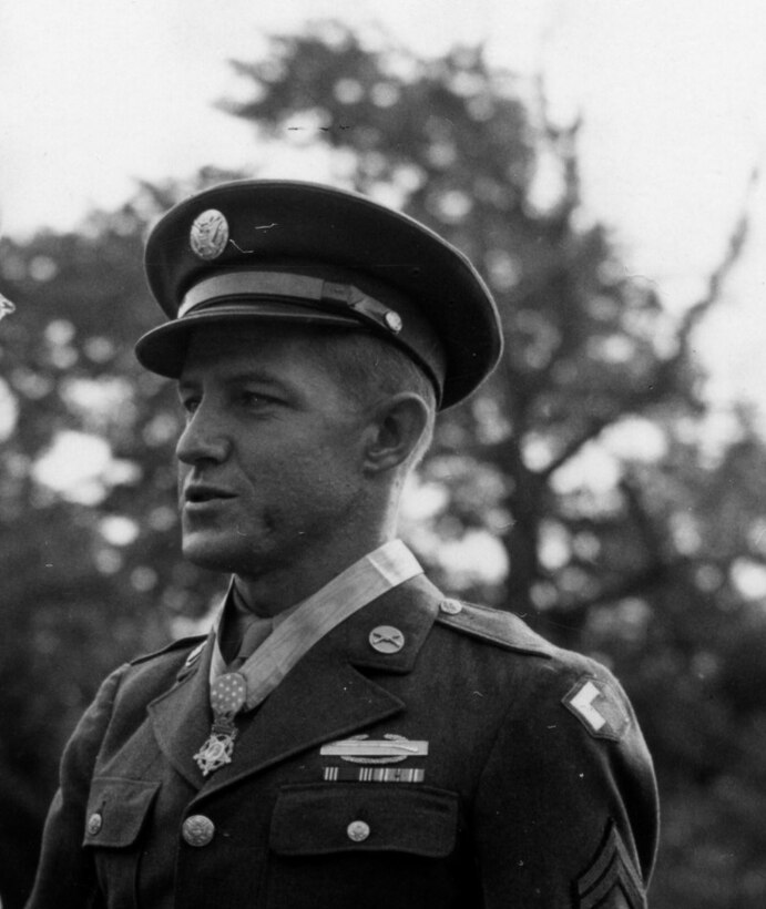 A service member in dress uniform and cap wears a medal around his neck.
