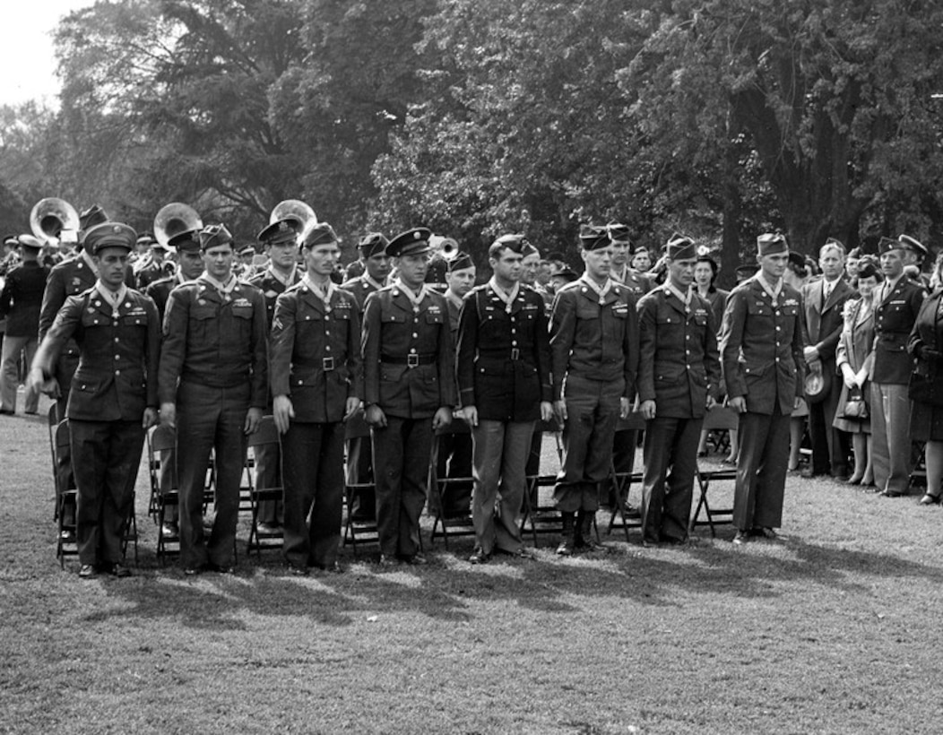 Two rows of service members stand at attention on a lawn. A band and an audience are in the background.