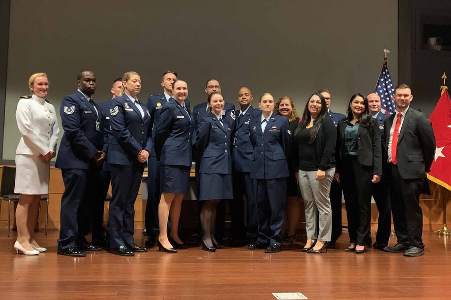 More than a dozen people, some in uniform and others in civilian attire, stand together on a stage.