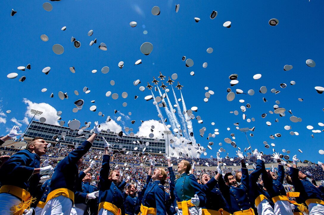 U.S. Air Force Academy graduates toss their hats in the air in a stadium as a formation of jets fly overhead.