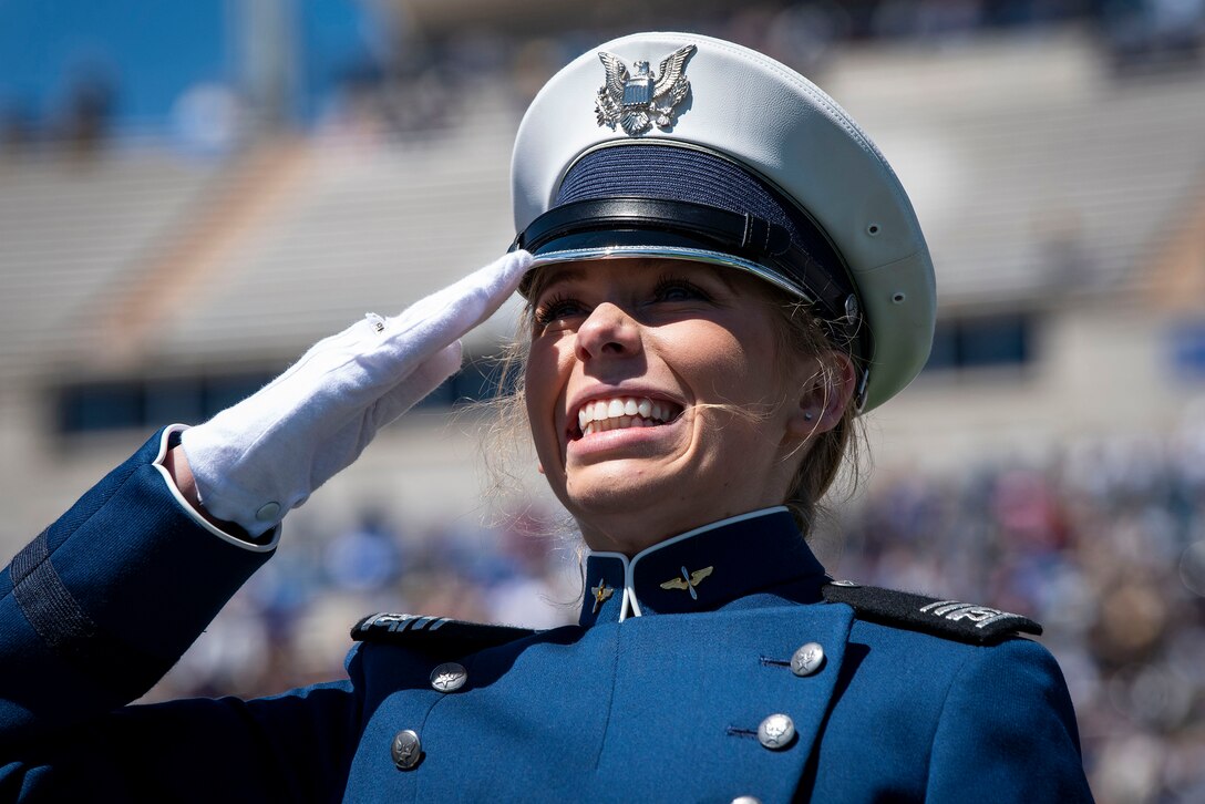 A U.S. Air Force Academy cadet salutes and smiles in a stadium.