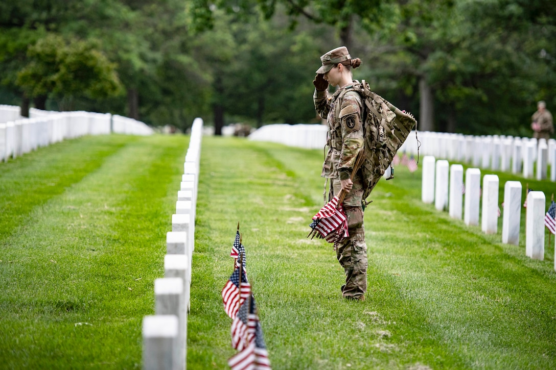 A service member holding small U.S. flags in one hand salutes in front of a cemetery headstone with the other.