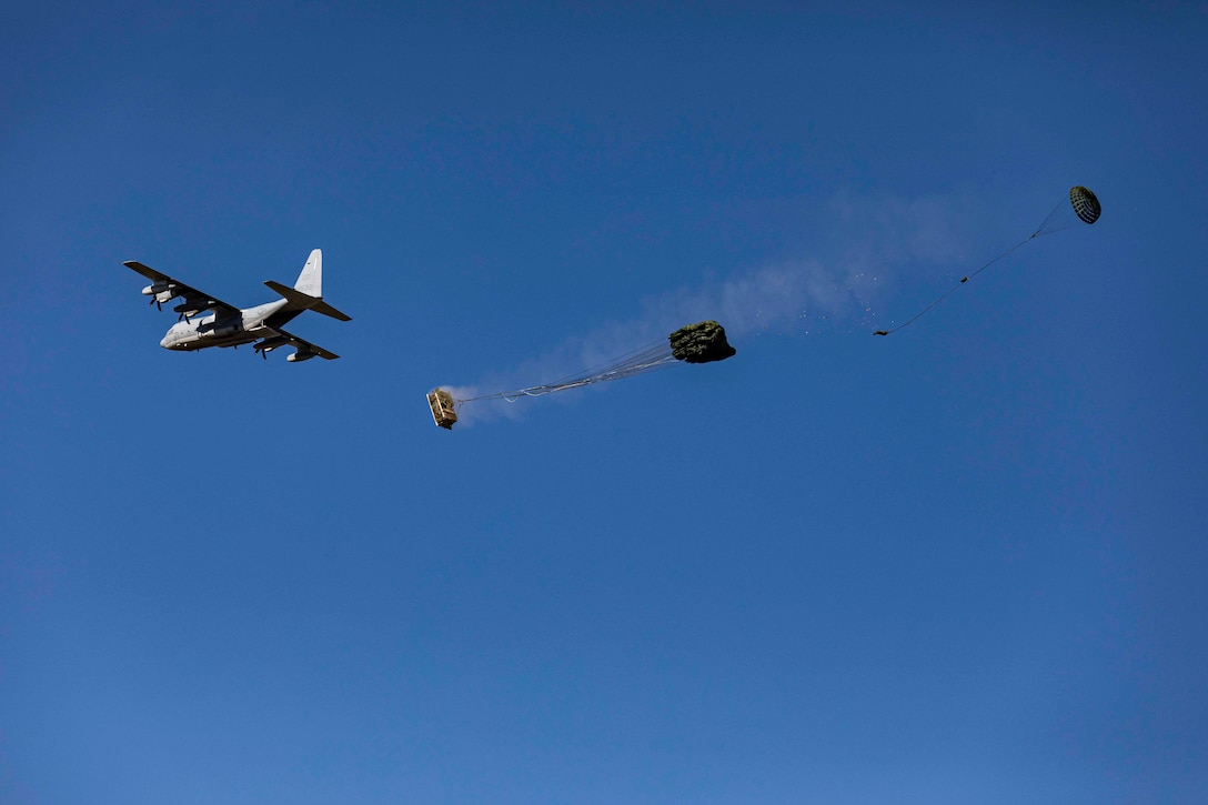 Two packages descend in the sky attached to parachutes as an aircraft flies in the background.