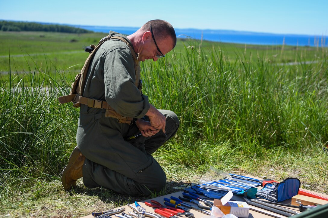 A service member kneels on the ground and manipulates tools.