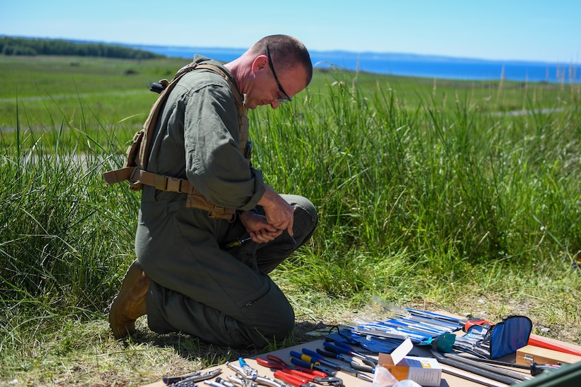 A service member kneels on the ground and manipulates tools.