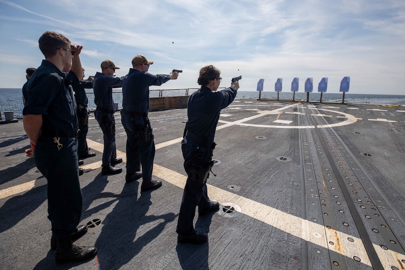 Service members on the deck of a ship fire handguns at targets.